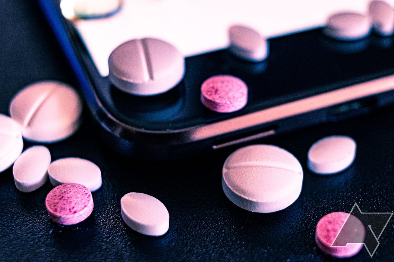 Μια κοντινή όψη της γωνίας μιας οθόνης smartphone με πολλά ροζ χάπια διάσπαρτα πάνω της.  Τα χάπια έχουν διαφορετικά σχήματα και μεγέθη και μερικά είναι μερικώς κομμένα στην άκρη της οθόνης.  Το φόντο είναι θολό, αλλά φαίνεται να είναι ανοιχτόχρωμο με κάποια υφή.