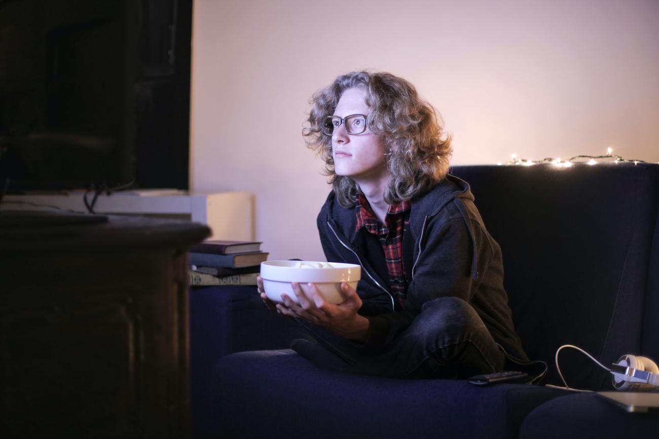 A man with with long hair and glasses holds a bowl of popcorn as he watches televsion. 