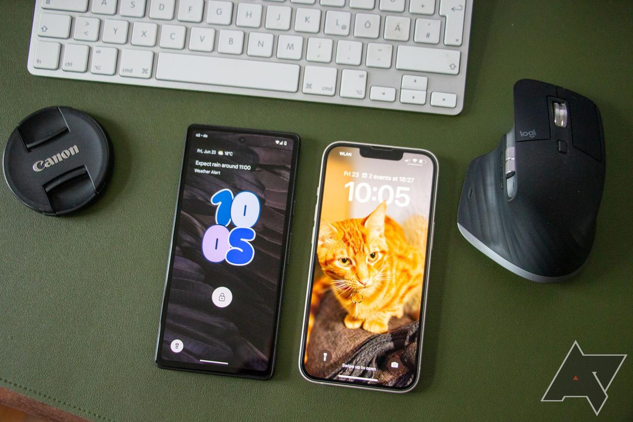 A Google Pixel 7a and an iPhone 13 lie next to each other on a green leather surface, with a Canon camera cap, an Apple Magic Keyboard, and a Logitech mouse artistically arranged around them