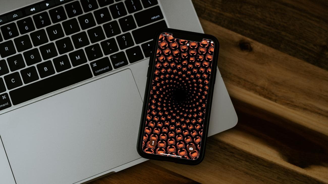 An iPhone on top of a Macbook laptop on a wooden table, with the iPhone displaying a spiral of crab emojis, which is one way you can edit your iOS 16 lock screen, if you