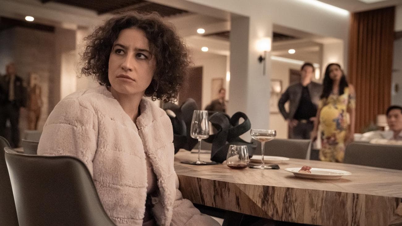 A woman with a fluffy jacket sits at a dinner party looking worried.