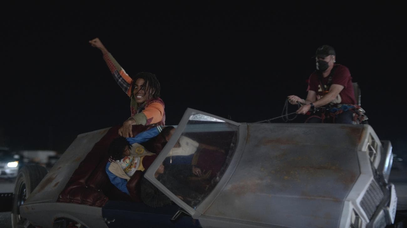 Ένας άντρας κρατιέται στο πλάι ενός μικροσκοπικού σκουριασμένου αυτοκινήτου με ρεαλιστικές ανθρώπινες κούκλες στα καθίσματα του, ενώ ένας άνδρας με κάμερα κινηματογραφεί.