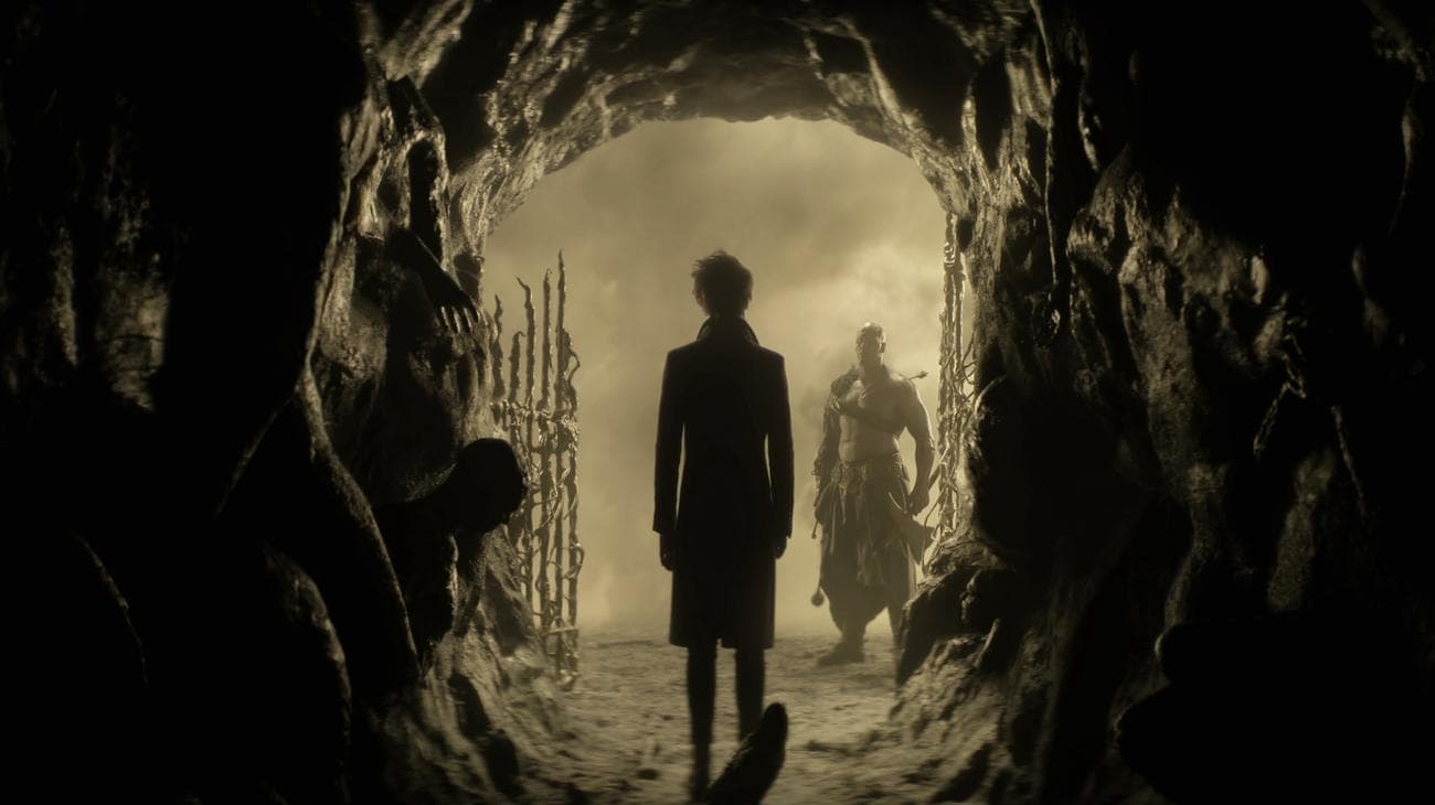 A man stands at the circular cave entrance to a realm, with a bodyguard awaiting.