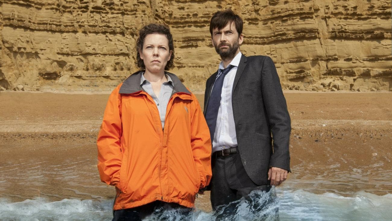 A woman in an orange jacket and a man in a suit jacket stand in the waves of a beach.