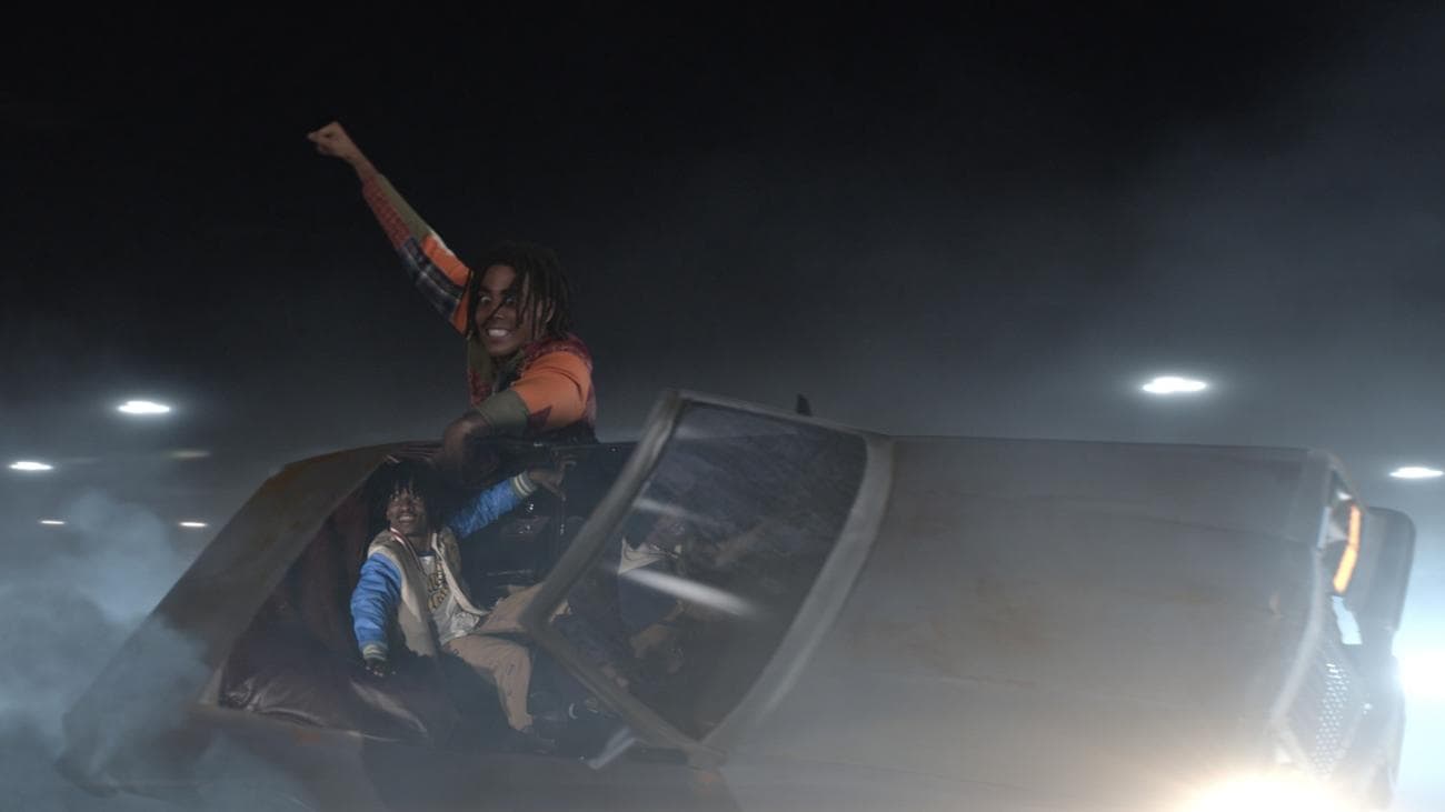 Ένας γίγαντας ύψους 13 ποδιών κρατιέται πάνω σε ένα αυτοκίνητο γεμάτο ενθουσιασμένους νέους καθώς κάνει έναν κύκλο γύρω από ένα πάρκινγκ.