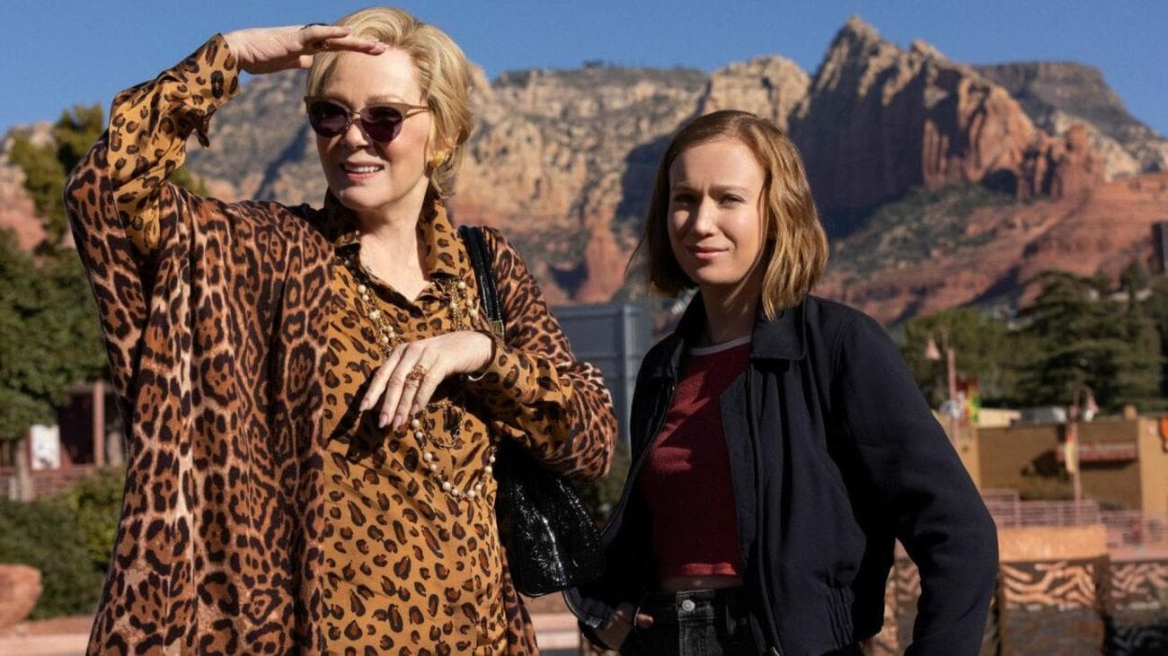 A woman in a leopard print shirt and a girl in a black jacket stand in front of a mountain.
