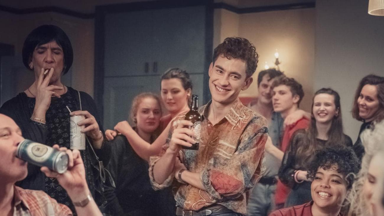 A young man stands with a beer smiling in the middle of a party.
