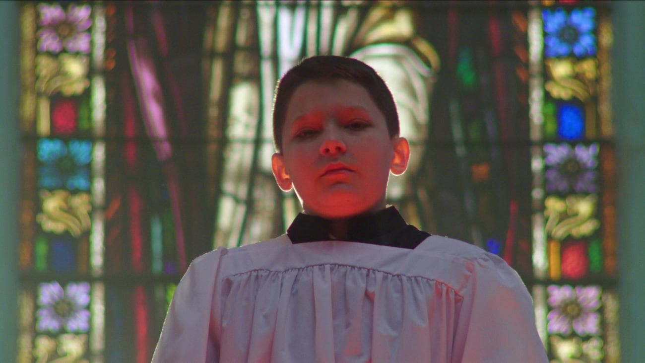 A choir boy stands in front of a stained glass window.
