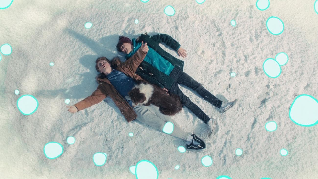 Two boys lie on snow with a dog, with animated snowflakes falling.