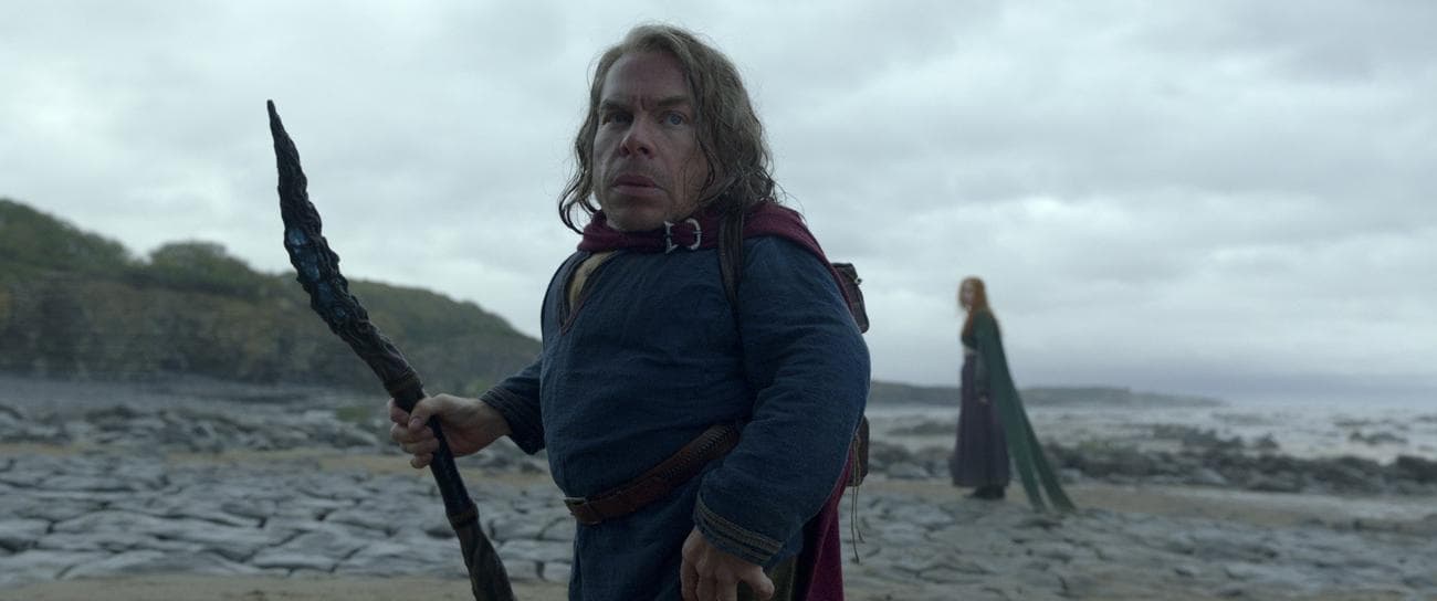 Ο Warwick Davis στο «Willow» κρατά ένα ραβδί, ενώ ένας άλλος χαρακτήρας στέκεται στο βάθος.