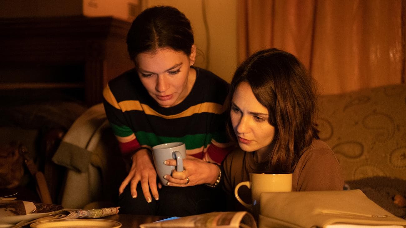Hannah Gross and Tuppence Middleton sit with cups of tea reading a phone on a coffee table in Albert Shin's Disappearance at Clifton Hill.