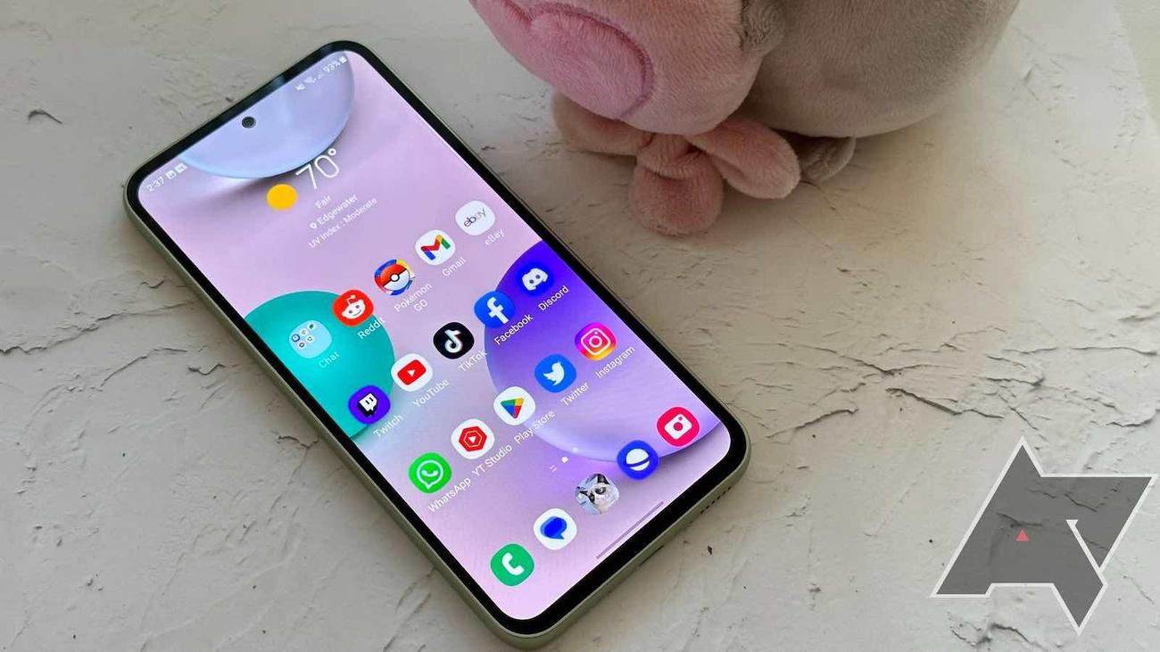 Ένα Samsung Galaxy A54 με φωτιζόμενη αρχική οθόνη στηρίζεται σε μια ανάγλυφη λευκή επιφάνεια.  Ένα ροζ βελούδινο αντικείμενο βρίσκεται στα δεξιά του. 