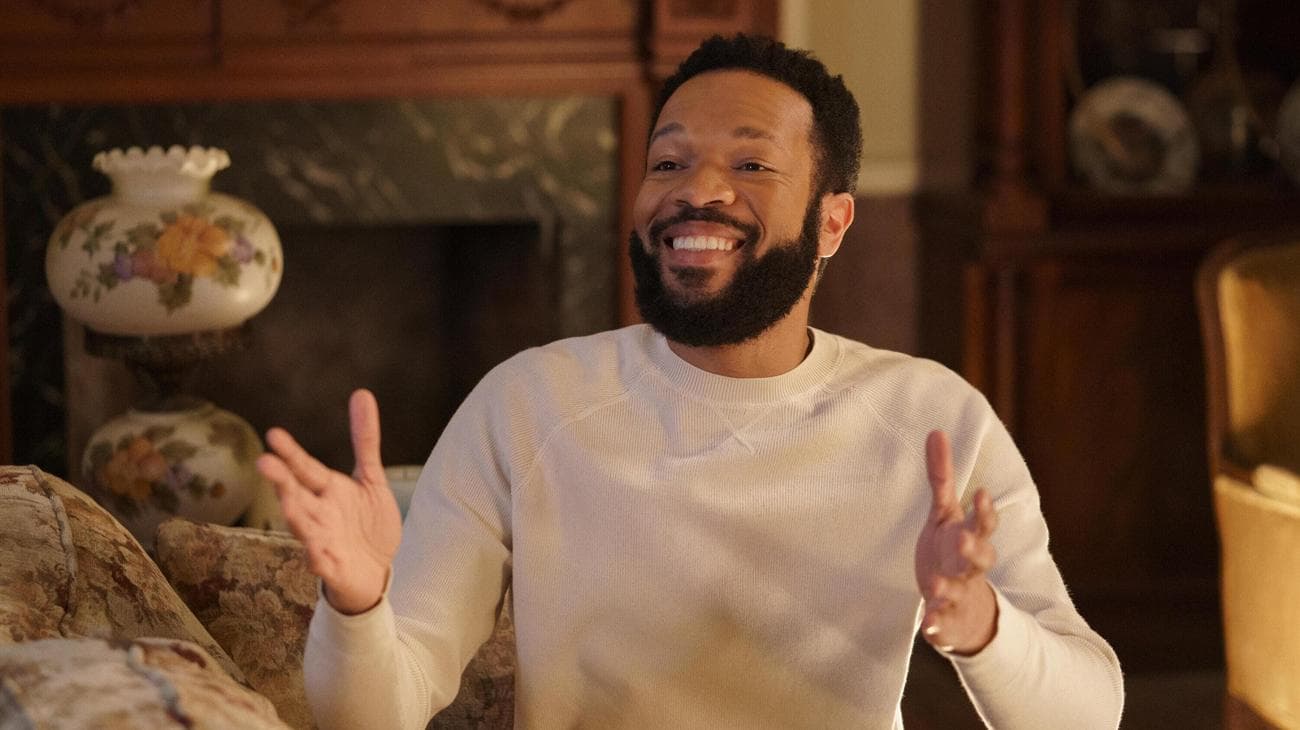 A young man wearing a white sweater laughs while sitting in a living room. 