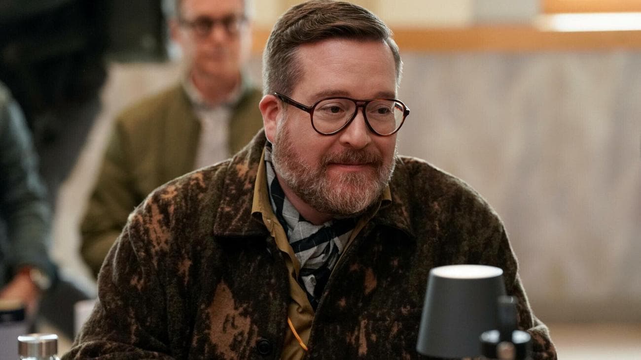 A man wearing glasses and a knitted sweater sits at a table read. 