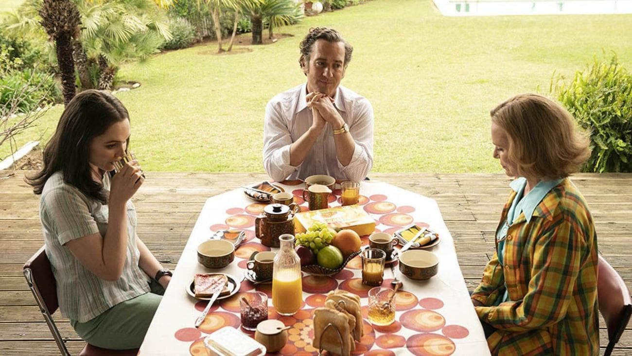 Three people sit awkwardly at an outdoor dining table in the TV show "Stonehouse."