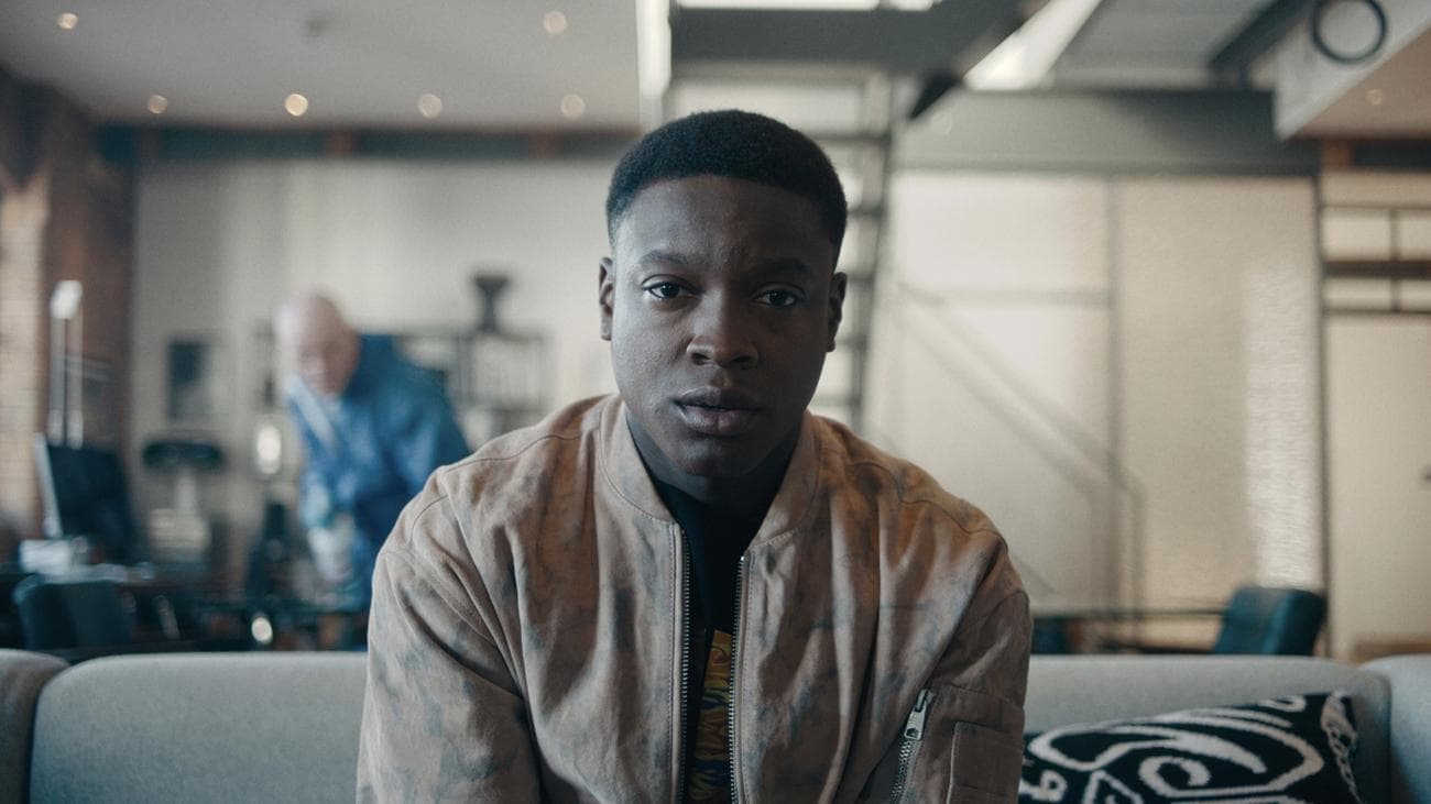 Ένας νεαρός άνδρας κάθεται φοβισμένος σε έναν καναπέ, ενώ ένας άντρας με μπλε πλαστικό καθαριστικό κρύβεται πίσω του.