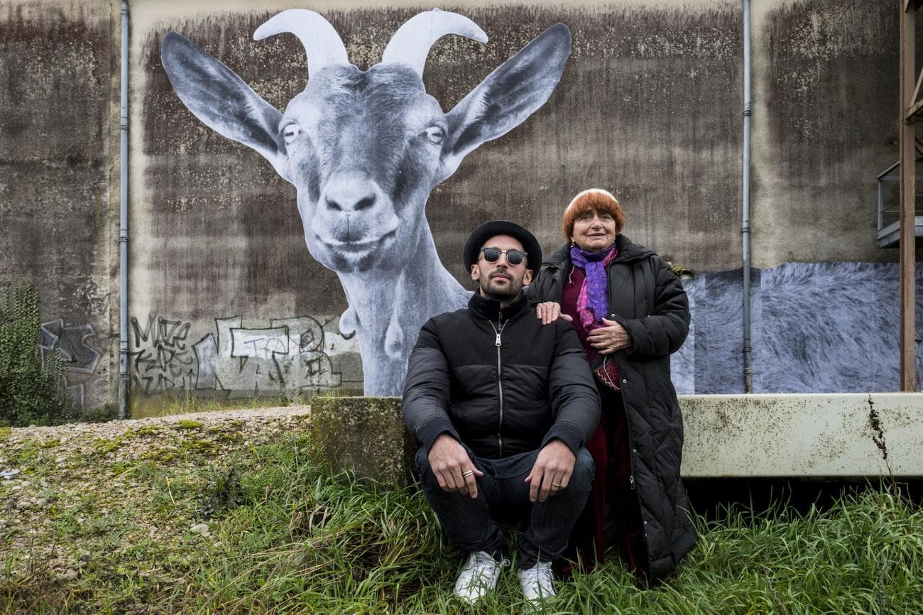 Agnes Varda and JR pose for a photograph in front of JR's artwork, a goat on a wall.