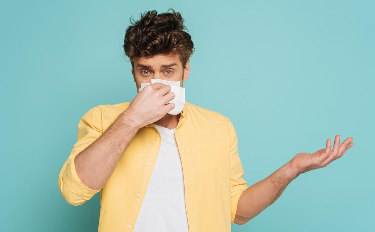 Person with short brown hair and bright yellow shirt on a light blue background holding tissue up to runny nose