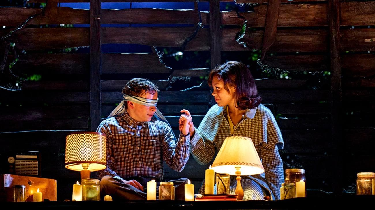 Σε "Stranger Things: The First Shadow" δύο έφηβοι κάθονται σε μια σοφίτα με κεριά, το αγόρι με δεμένα τα μάτια.