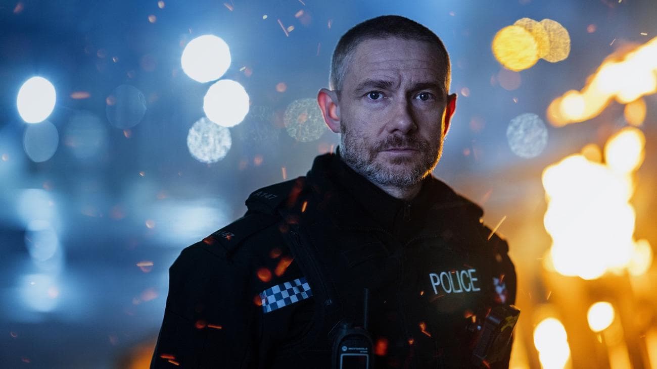 A cop stands on a street in the night as a building burns behind him.