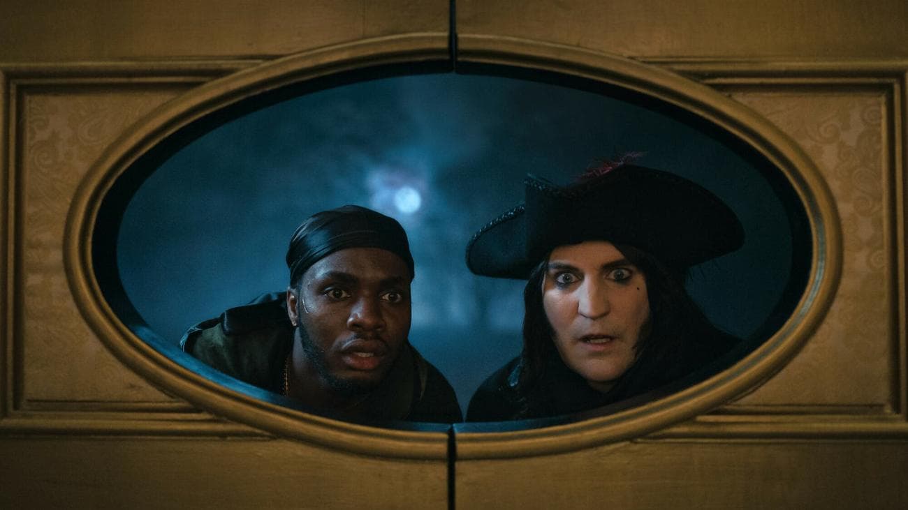 Η Duayne Boachie και ο Noel Fielding με ρούχα του 18ου αιώνα κοιτάζουν μέσα από ένα στρογγυλό παράθυρο.