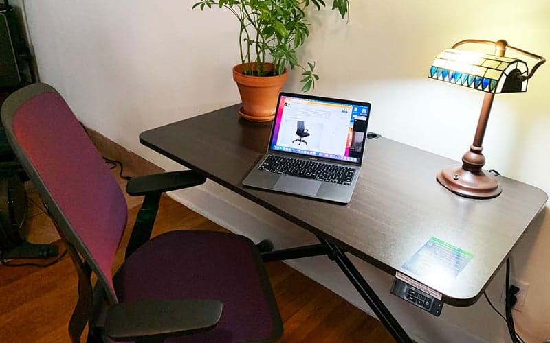 The mesh Steelcase Karman Office Chair is facing a brown standing desk with an open laptop, plant, and a lamp on top.