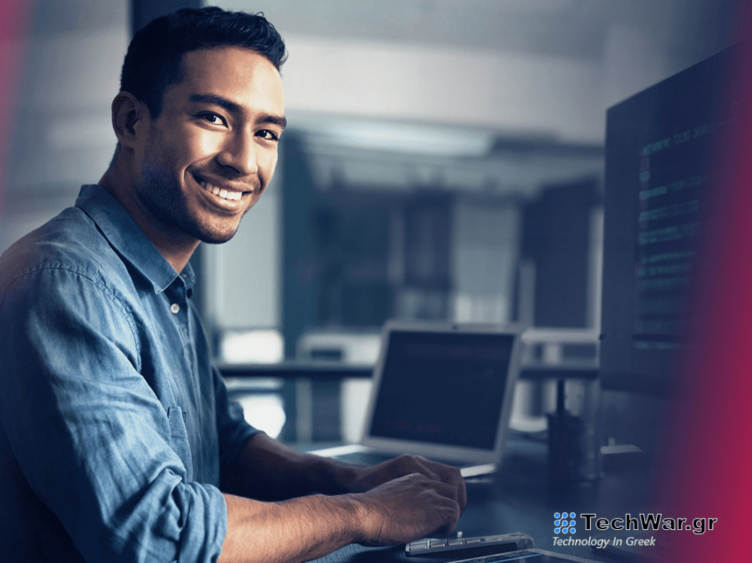 A person looking at the camera and smiling while typing on a computer.