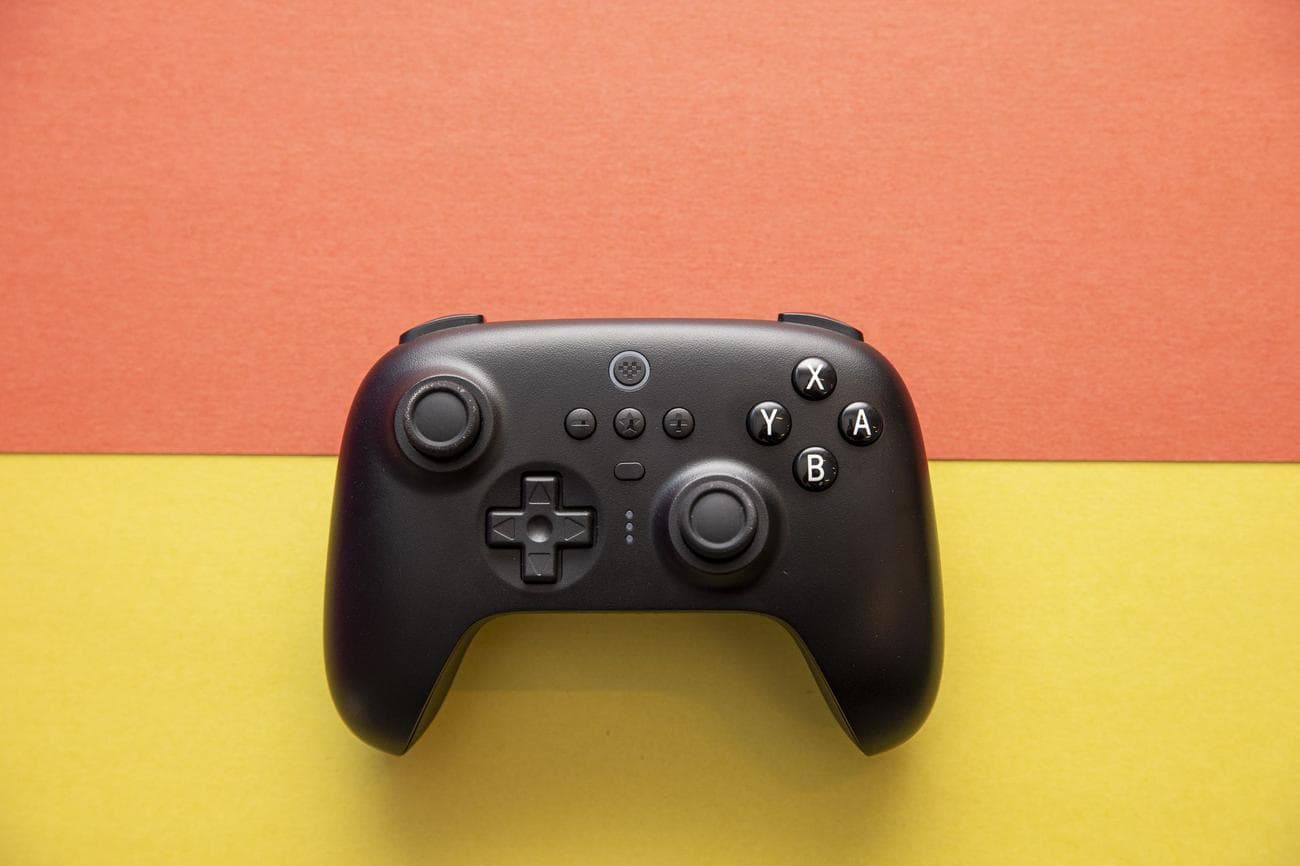 Top-down shot of 8BitDo Ultimate Controller on an orange and yellow background. The controller has an asymmetric joystick design, like an Xbox or Switch Pro controller, and it's black, with white legends on the A B X Y buttons.