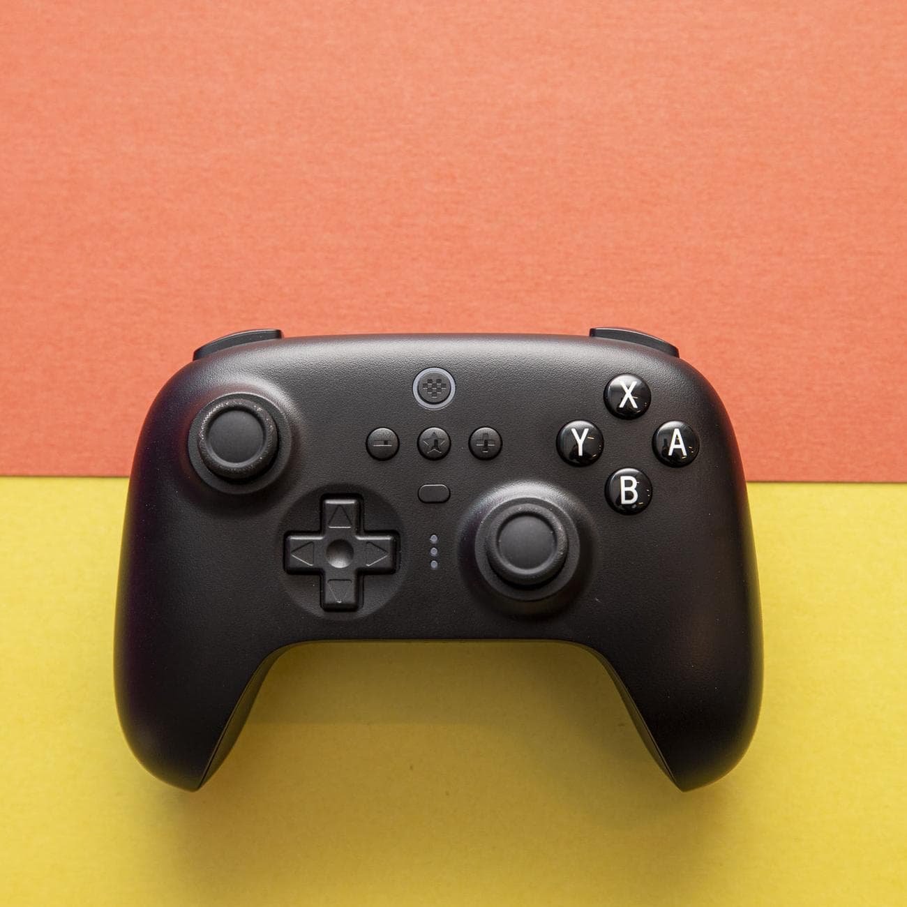Top-down shot of 8BitDo Ultimate Controller on an orange and yellow background. The controller has an asymmetric joystick design, like an Xbox or Switch Pro controller, and it's black, with white legends on the A B X Y buttons.