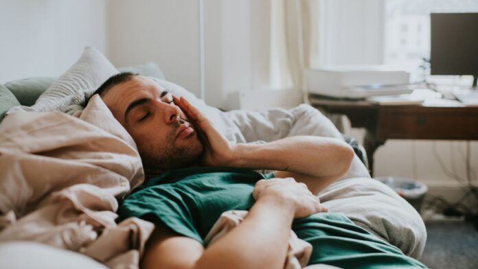 A sleep deprived man in a green shirt falls asleep for a second on his sofa, having what experts refer to as a microsleep because he