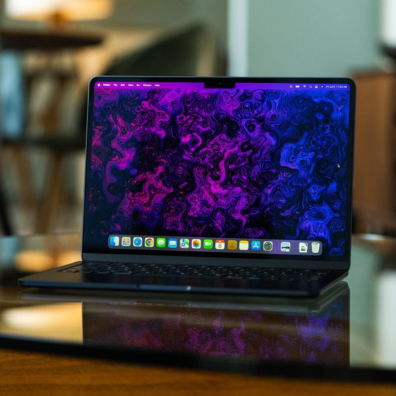 The M2 MacBook Air is opened, facing the camera. Its display is on, showcasing a psychedelic purple and black wallpaper created by The Verge's art and illustration team.