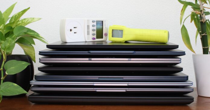 A stack of laptops on top of a brown table, flanked by two green plants, against a white wall.